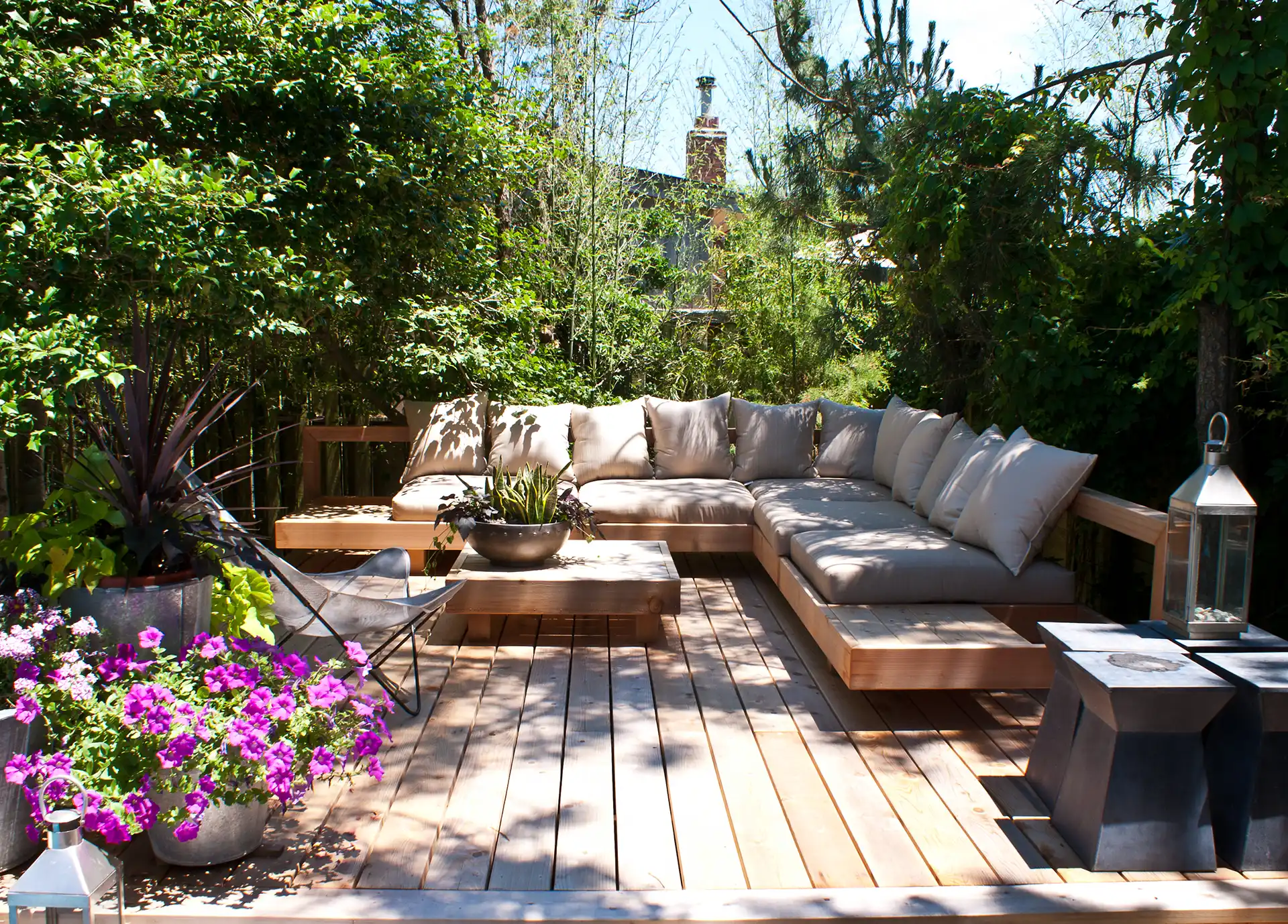 Helle Holzterrasse mit großzügiger Lounge-Ecke und Gartenmöbeln, umgeben von üppigem Grün und Blütenpflanzen.